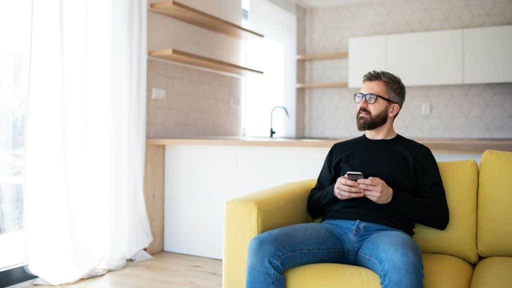 A thoughtful, bearded man with glasses, wearing a black top and blue jeans, sits on a bright yellow couch, holding a smartphone.