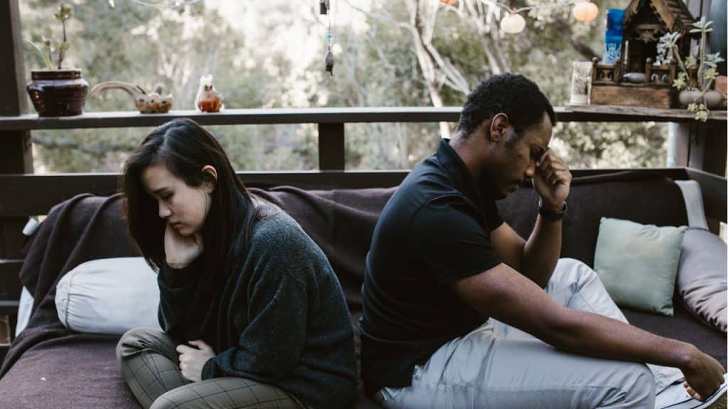 A man and a woman sit back-to-back on a porch sofa, both looking upset and resting their heads on their hands.
