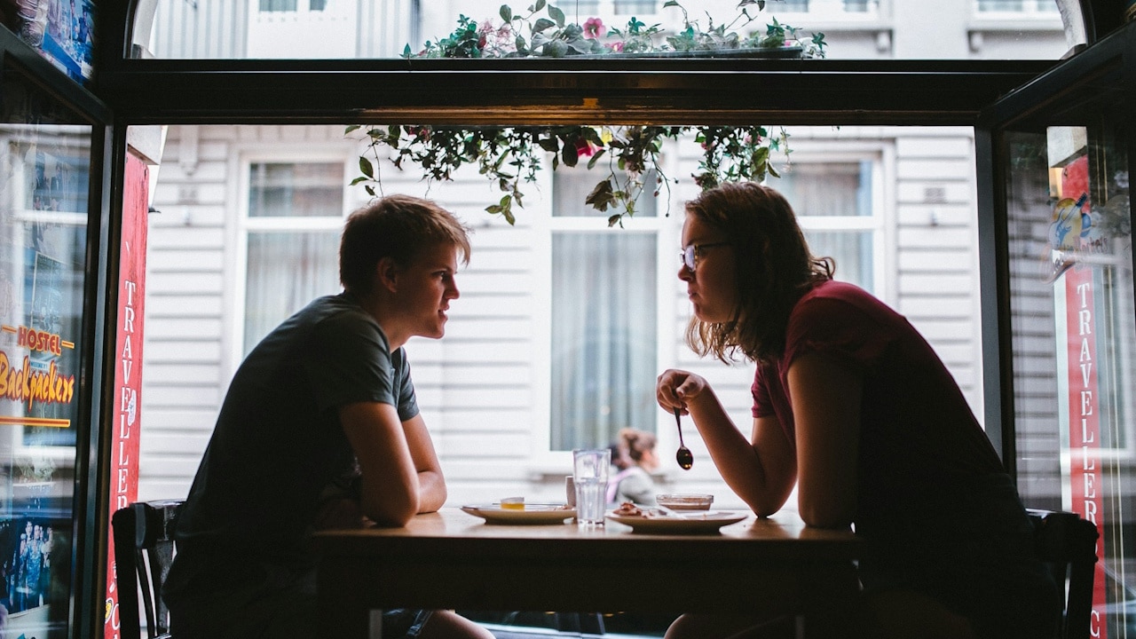 A young man and woman sit opposite each other at a table, looking into each other's eyes, framed by a dark cafe window.