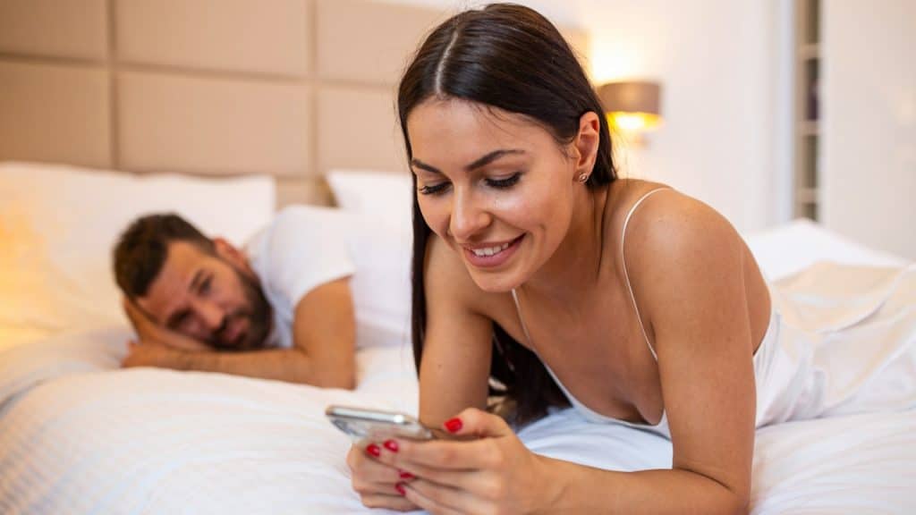 A smiling woman is lying on her stomach in bed, looking at her smartphone, while a man lies behind her in the background.