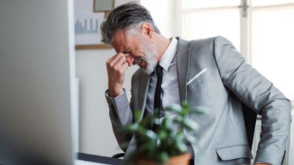 Middle-aged businessman in a grey suit holding his head in distress at his office desk.