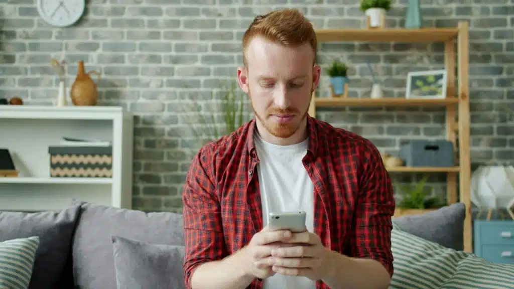 Red-haired man in a plaid shirt intently looking down at his smartphone on a grey couch.