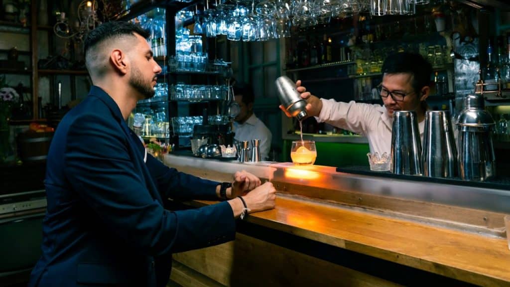 Two men at a dimly lit bar, one is shaking a cocktail for the other.
