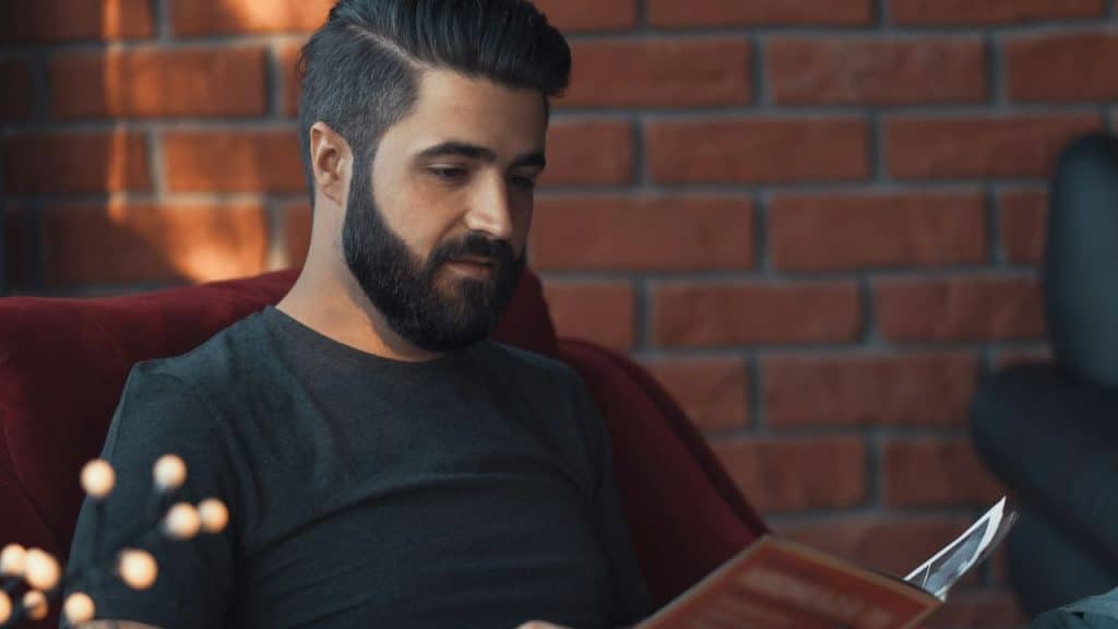 Bearded man in a dark shirt reading a book or magazine against a brick wall background.
