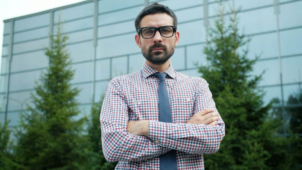 Man in glasses and a plaid shirt with a tie standing with his arms crossed outdoors.