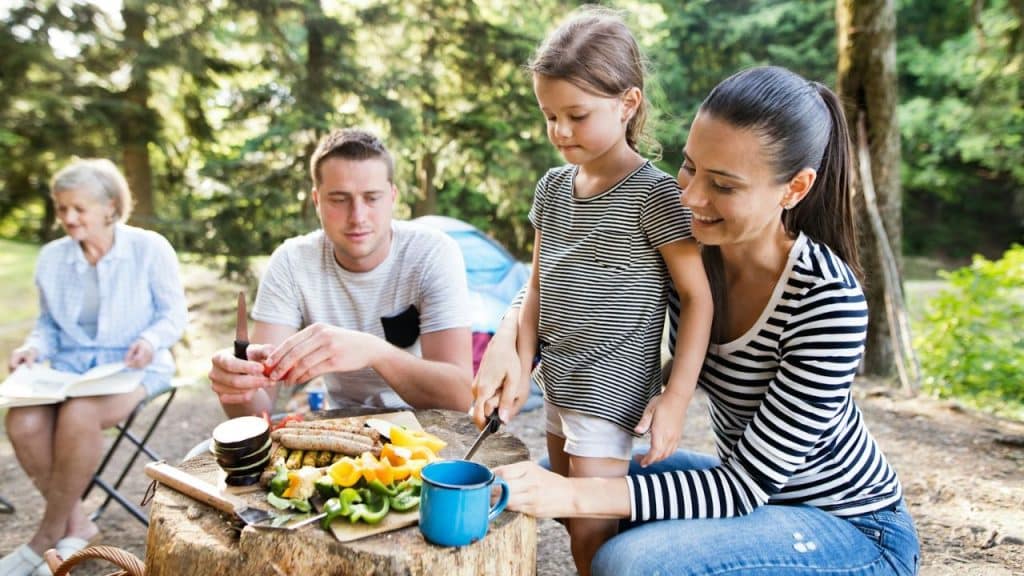 Family enjoying a picnic outdoors with food on a tree stump, with an older woman in the background.