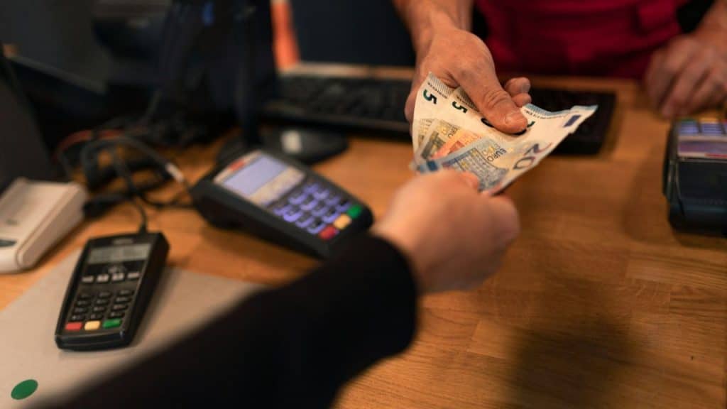 Close-up of hands exchanging Euro banknotes over a counter with payment terminals.