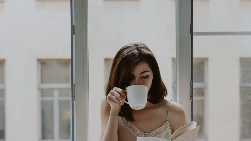 A woman sipping coffee and reading by a window.