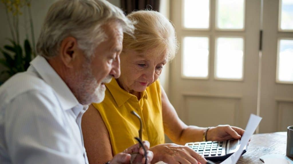 Elderly woman in yellow pointing at a document while a man watches, indoors.