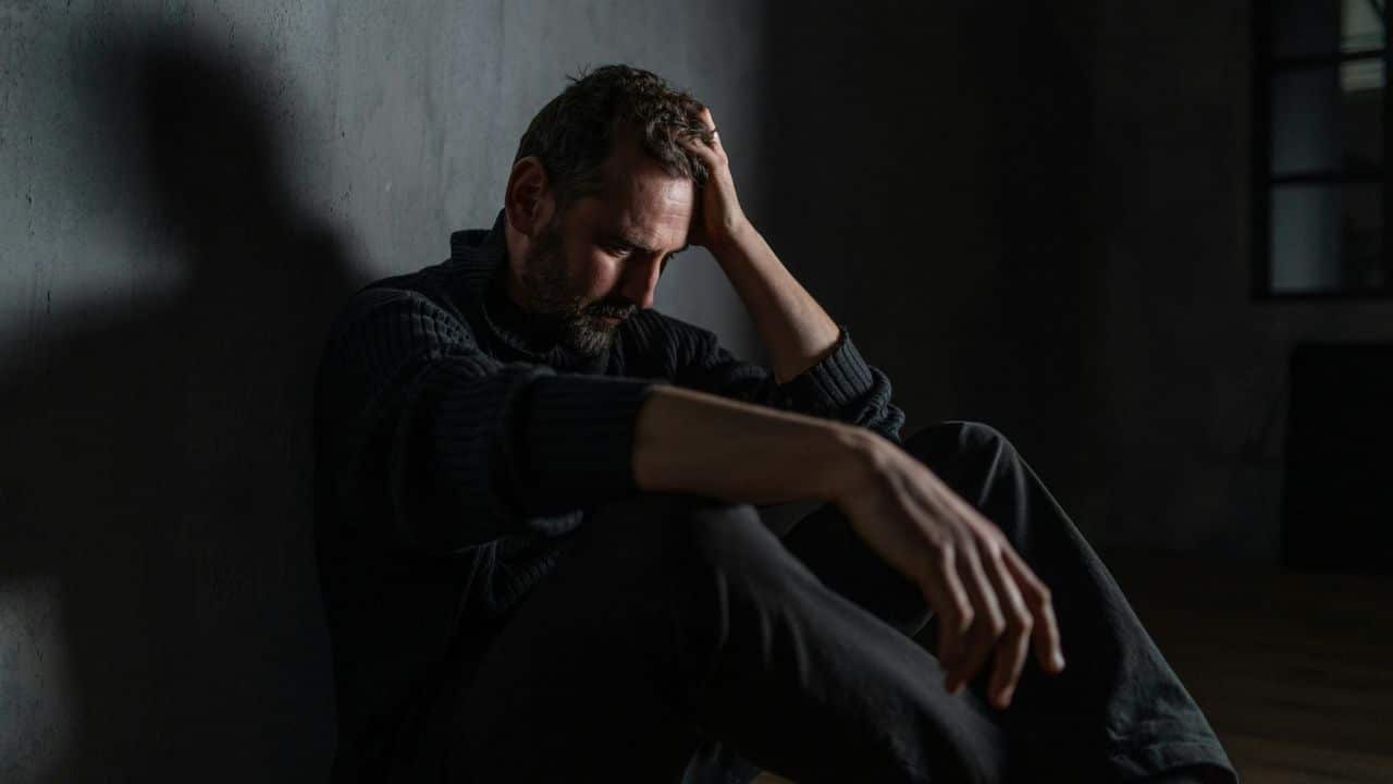 Distressed man sitting alone on the floor in a dimly lit room with hand on head.