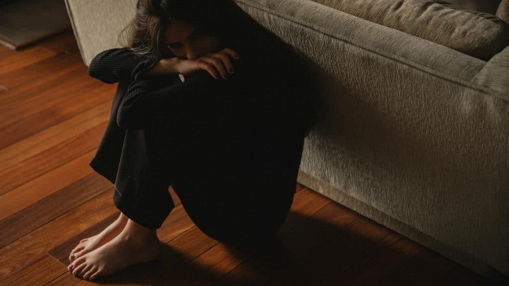 A person sits barefoot on the floor, leaning against a couch in dim light.