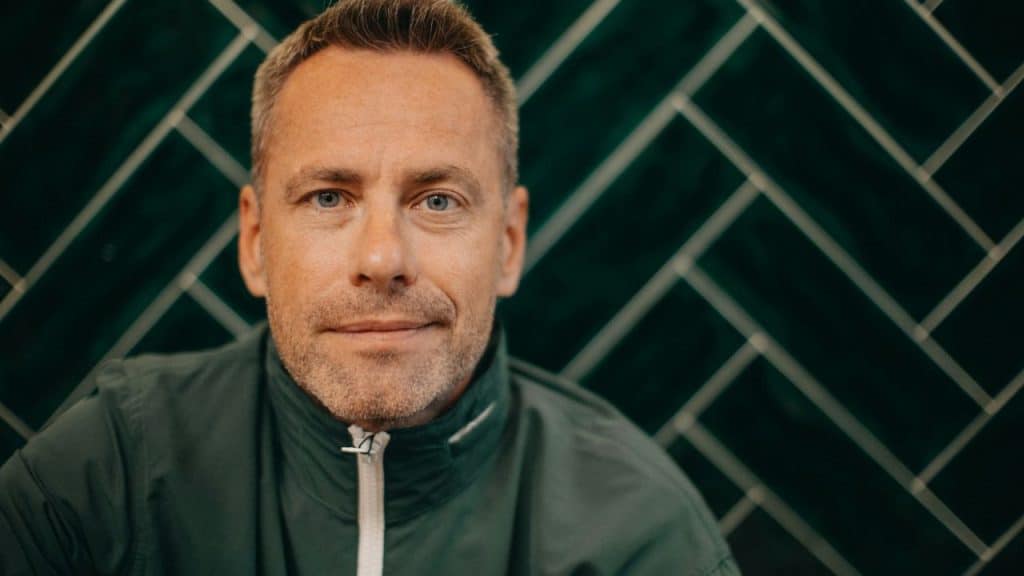 Close-up portrait of a man with blue eyes against a dark green tile wall.