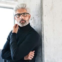 Man with gray hair and glasses in black turtleneck leans against a concrete column.