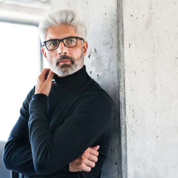 Man with gray hair and glasses in black turtleneck leans against a concrete column.