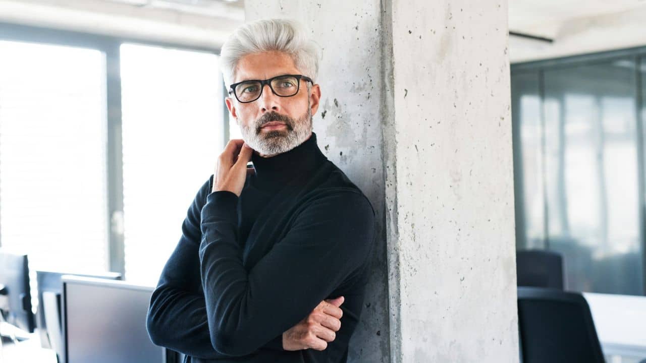 Man with gray hair and glasses in black turtleneck leans against a concrete column.