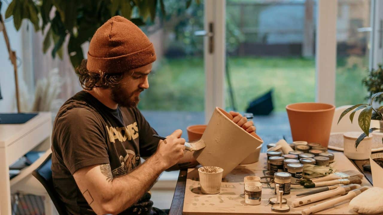 Bearded man in a knit cap painting a terracotta pot at a table full of art supplies.