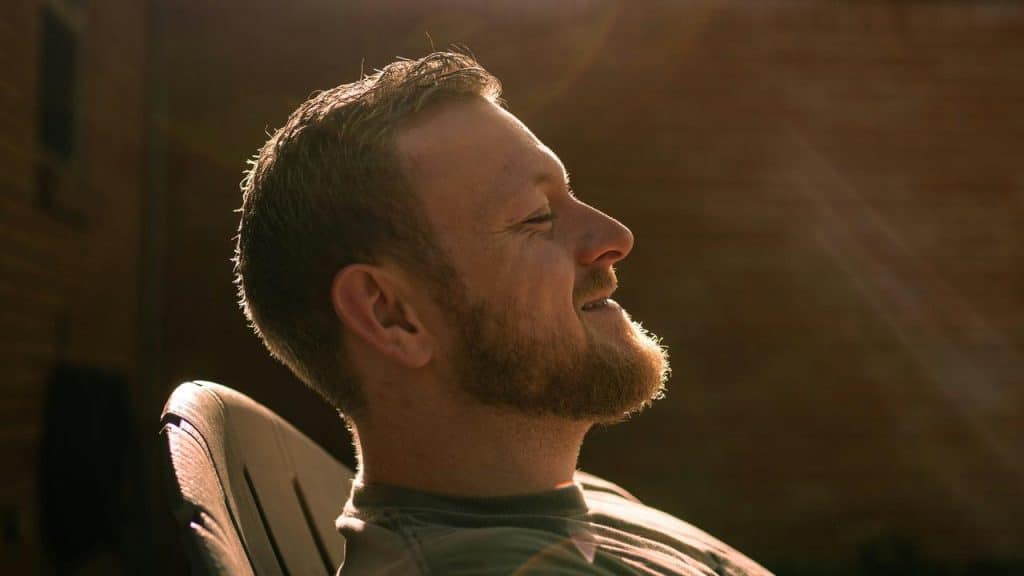 A man with a beard relaxes in a chair outdoors, smiling peacefully in warm sunlight.