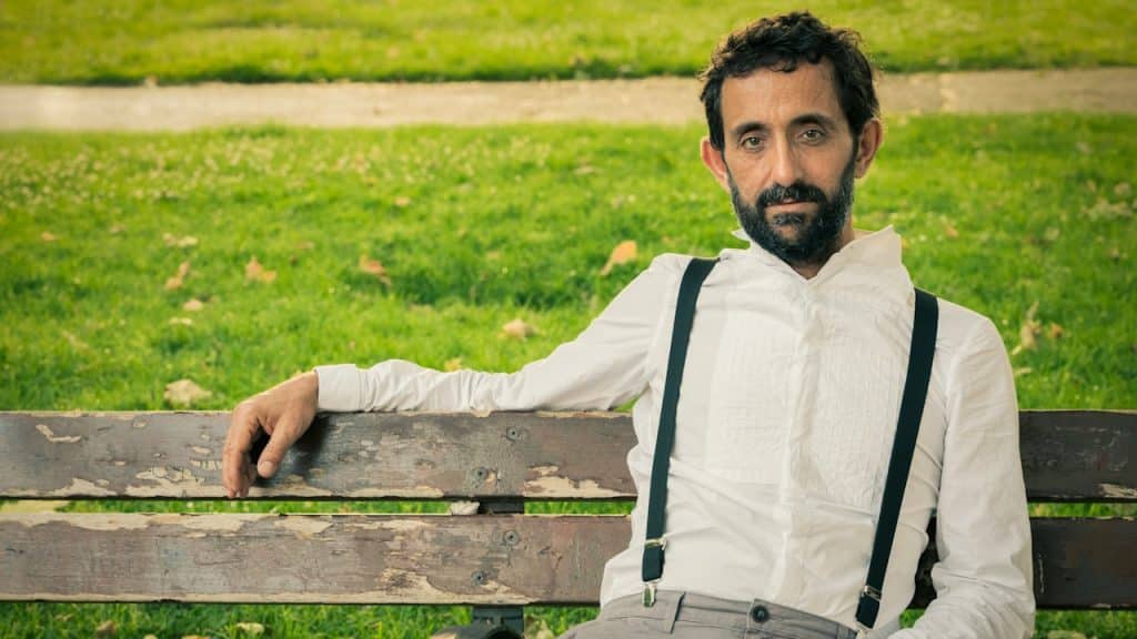 A bearded man wearing suspenders and sitting on a bench at the park.
