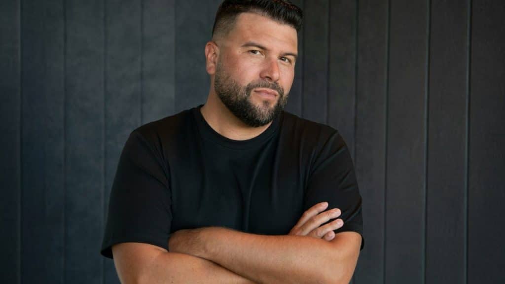 Bearded man in a black t-shirt standing with arms crossed against a dark wall.