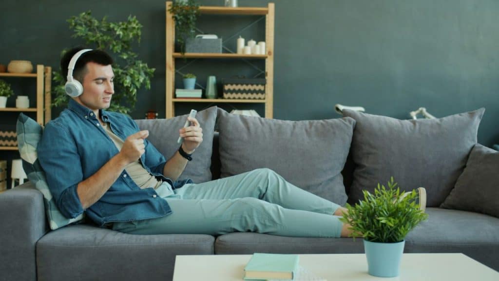 Young man relaxing on a gray sofa with headphones and looking at his phone.