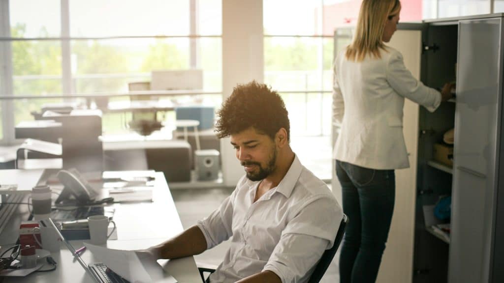 A man is working on the laptop while a woman stands behind him rifling through a cabinet.