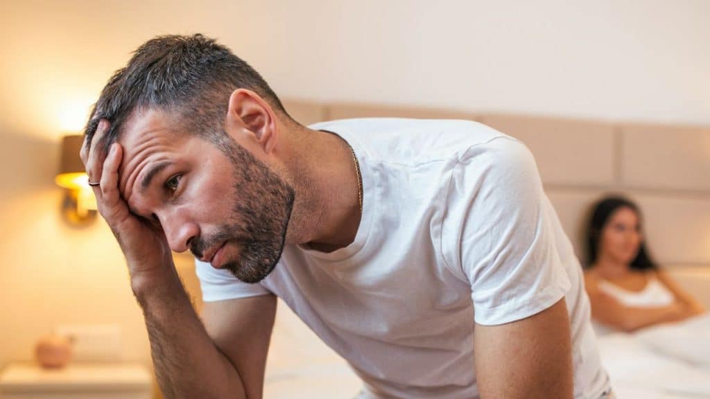 A distressed man sits in the foreground with his hand on his head; a woman is out of focus in the background.