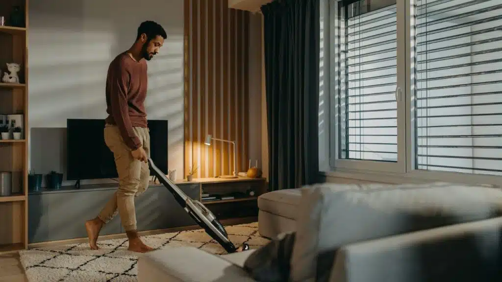 Barefoot man vacuuming a rug in a sunlit living room with blinds.