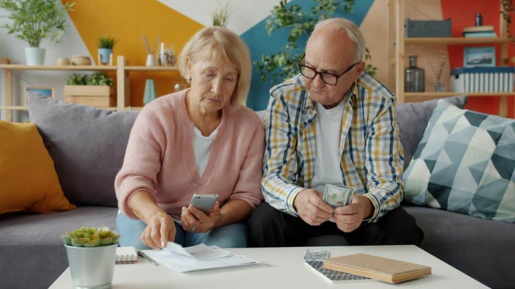 Elderly couple reviewing documents, the man counting money, the woman holding a smartphone.