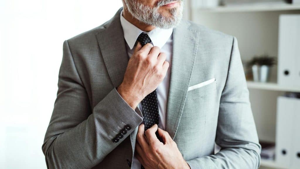 A man in a gray suit adjusting his black tie.
