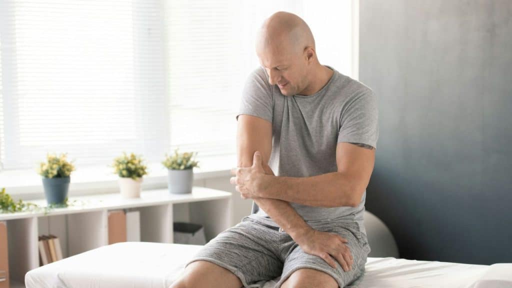 Bald man in a grey t-shirt and shorts sitting on a bench holding his elbow in pain.