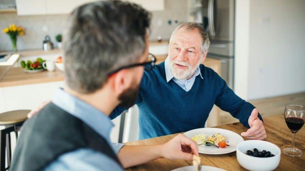 Two men of different generations eating and talking animatedly at a wooden kitchen table.