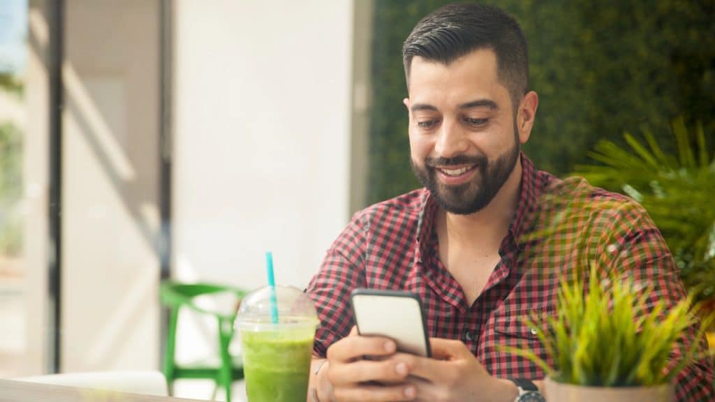 Smiling man in a checkered shirt checking his phone with a green smoothie nearby.