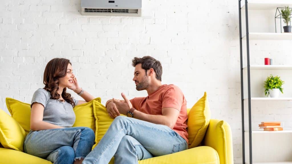 A couple sitting on a yellow couch talking in a bright room with an air conditioner.