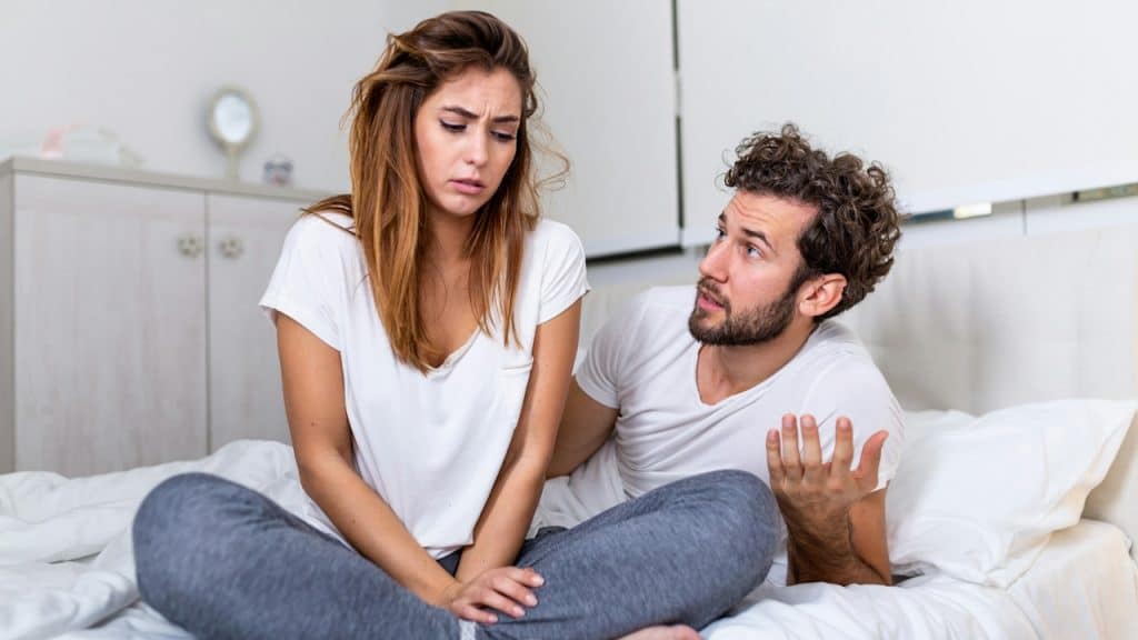 A lightly bearded man talking to a woman while they sit on a bed.