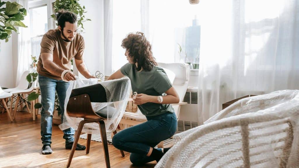 A man and woman wrapping a chair with bubble wrap in a bright room.
