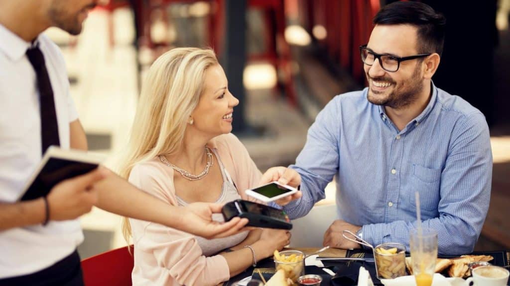 A man pays a restaurant bill using a phone while a smiling woman sits next to him.
