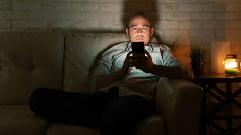 A man sits on a couch in a dark room, illuminated by his smartphone screen.
