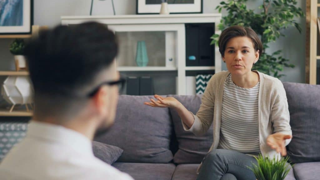 A woman sits on a couch, gesturing with her hands while speaking to a man facing away.