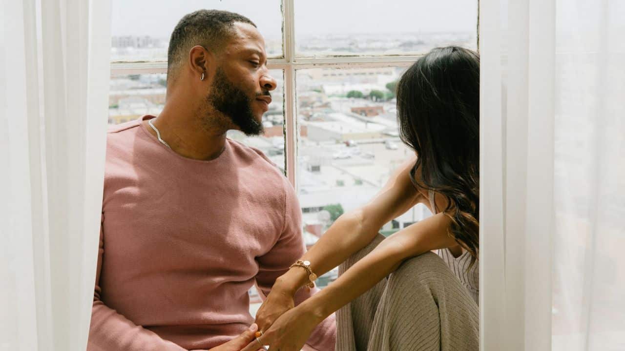 A man and woman sit by a large window overlooking a city, holding hands.