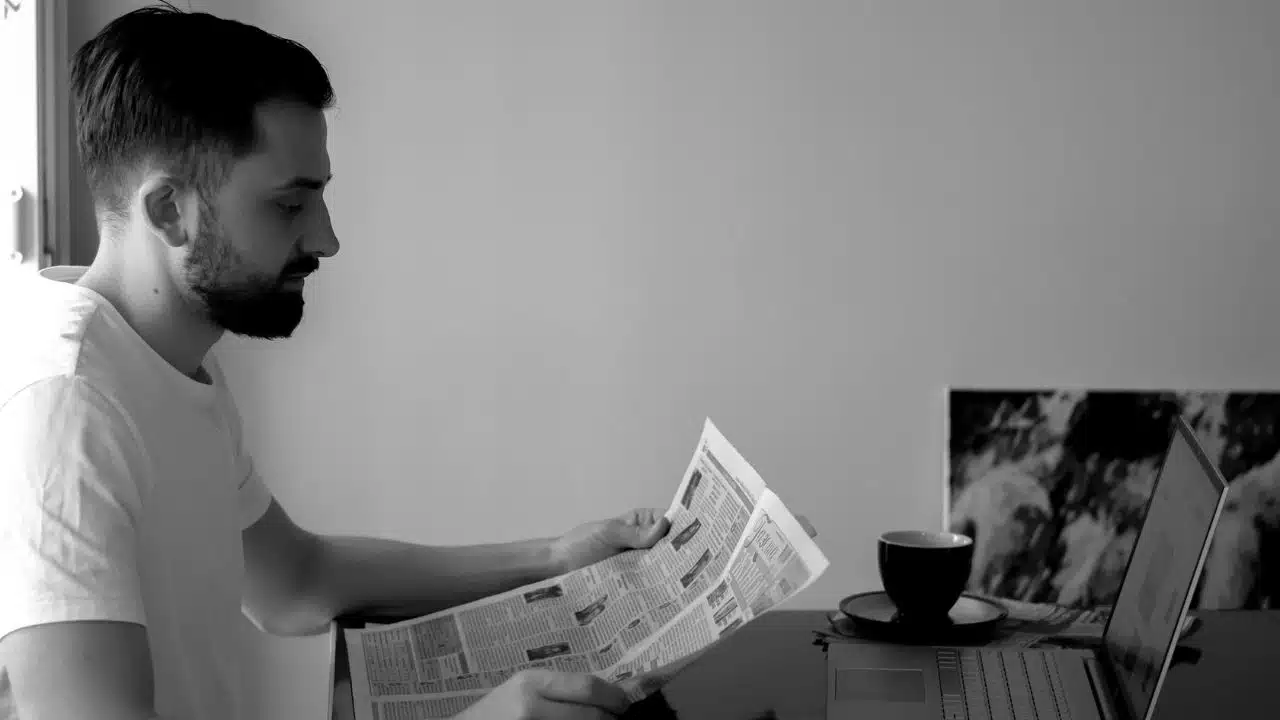 Black and white photo of a man reading a newspaper next to a laptop and coffee cup.