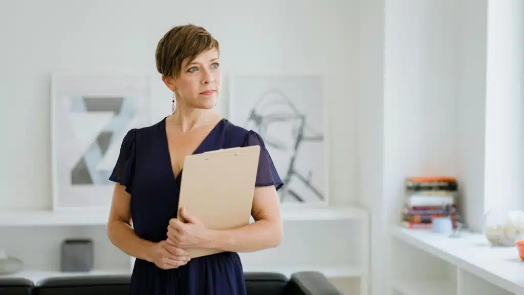 A woman holding a clipboard stands in a bright room, looking thoughtfully to the side.