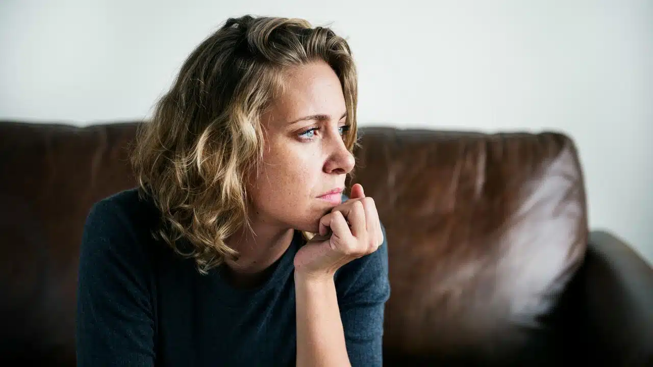 A woman sits on a couch looking thoughtful and concerned.