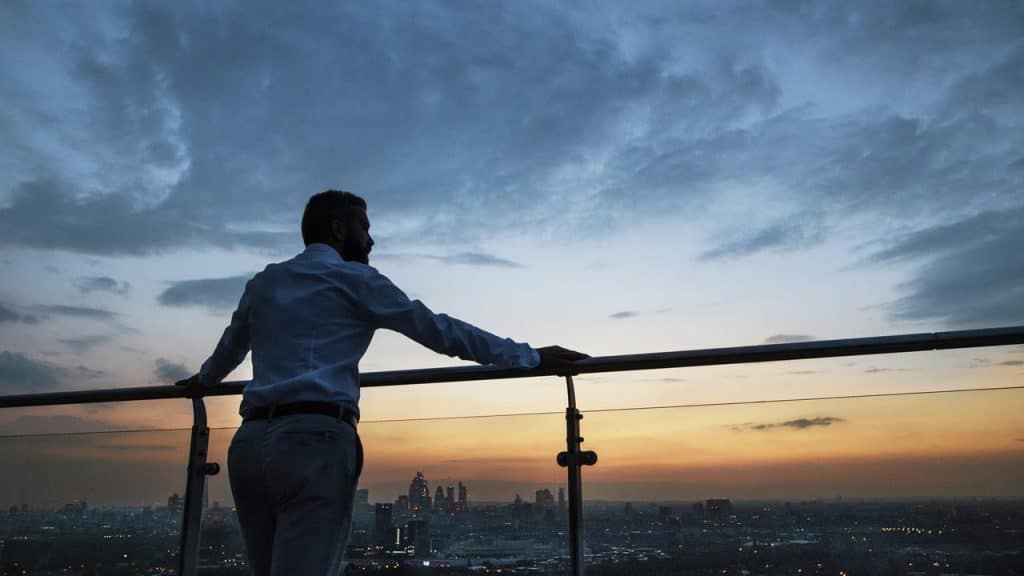 A man stands on a rooftop terrace looking at a city skyline during sunset.