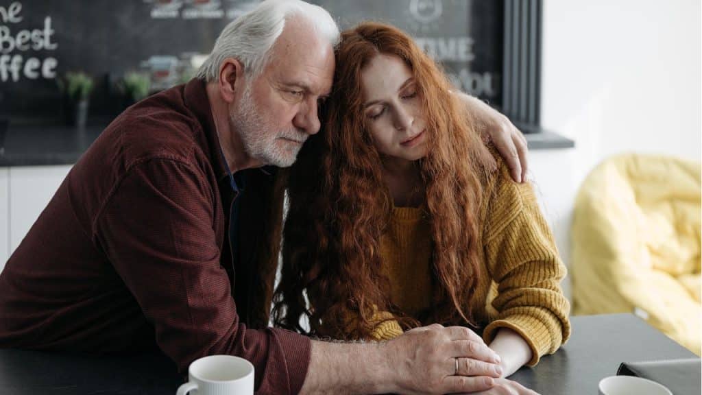 Older man comforting a younger, red-haired woman by putting his arm around her and holding her hand.