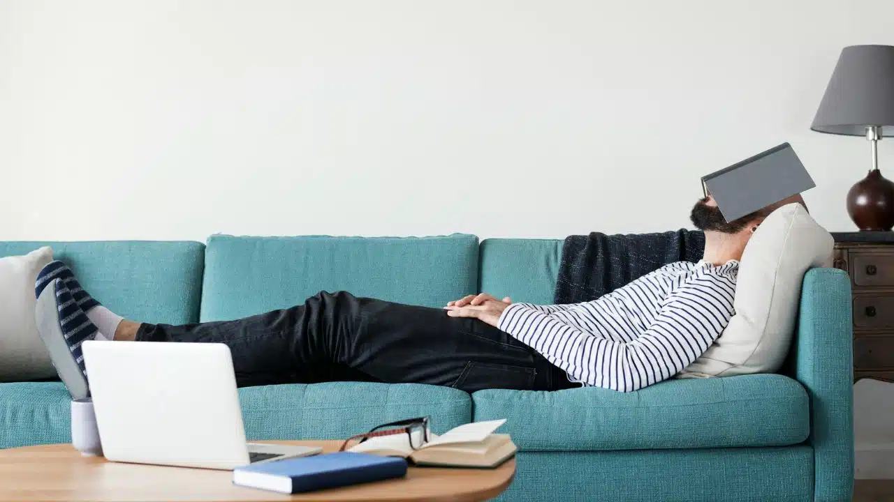 A man is lying on a turquoise couch with a tablet covering his face.