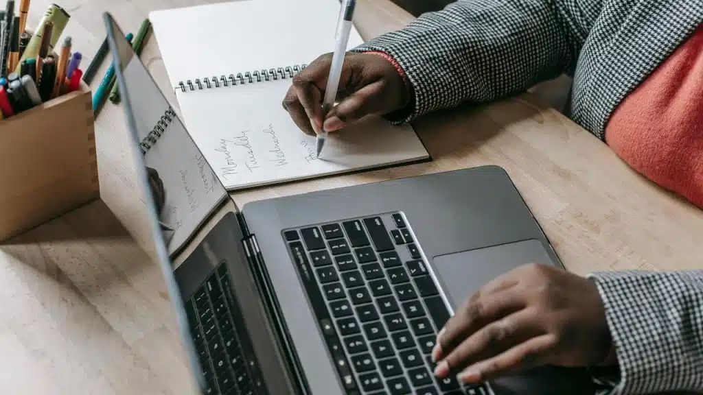 A person writing in a notebook while working on a laptop.
