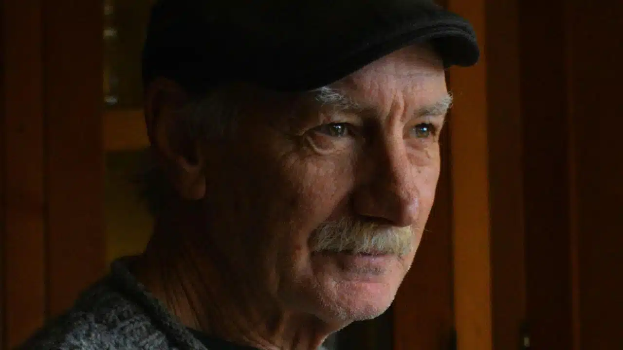 An older man with a mustache and a black cap is looking to the side.
