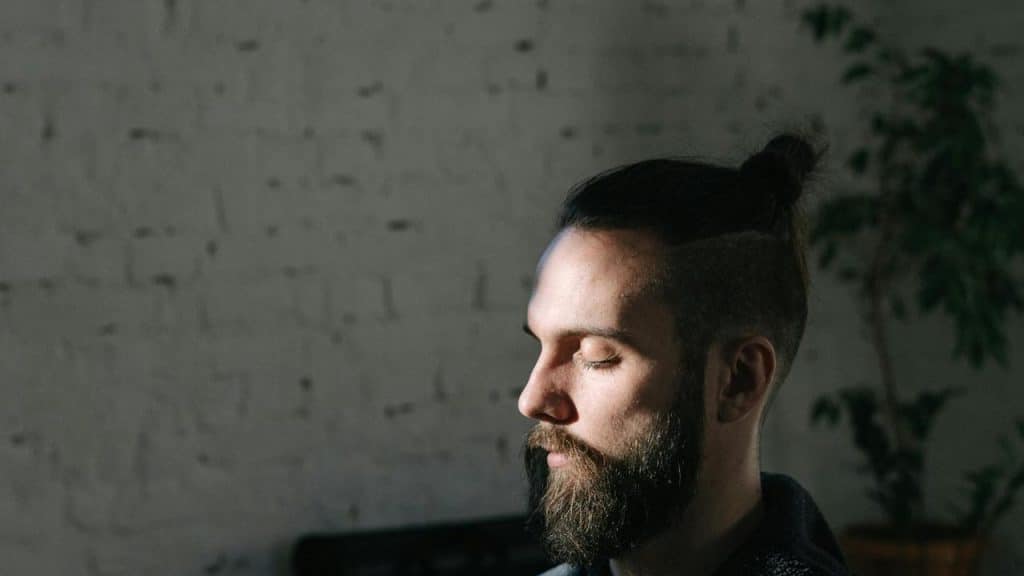 A bearded man with closed eyes sitting in partial sunlight indoors.