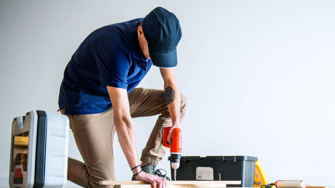 A man kneeling on the floor is using a power drill on a piece of wood.
