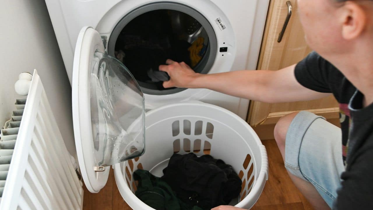 A person kneels, loading dark laundry into a washing machine from a white basket.